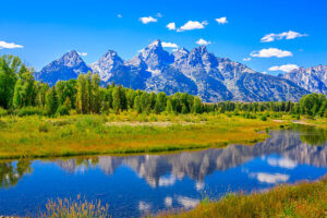 Stunning panoramic view of Jackson Lake in Wyoming, nestled at the foot of the majestic Teton Range. Snow-capped peaks reflect in the crystal-clear water, surrounded by lush green forests.