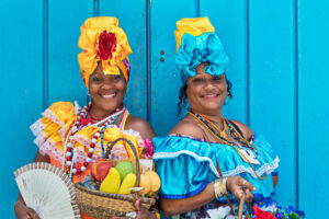 Portrait of happy Cuban women standing against blue wooden wall. Smiling mature women are in traditional dresses. They are with fruit basket and hand fan.